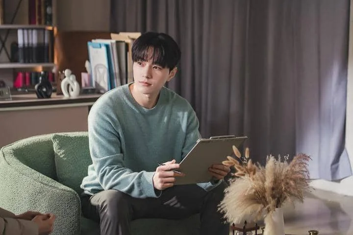 Seorang pria mengenakan sweater biru muda duduk di sofa hijau sambil memegang clipboard di ruangan dengan pencahayaan lembut.