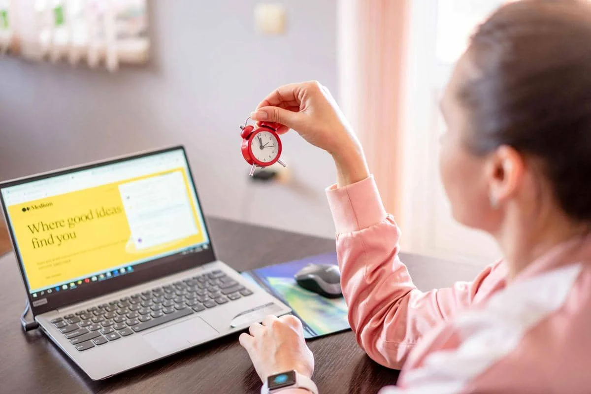 Seorang perempuan mengenakan pakaian merah muda memegang jam kecil berwarna merah di depan laptop yang menampilkan laman berwarna kuning.