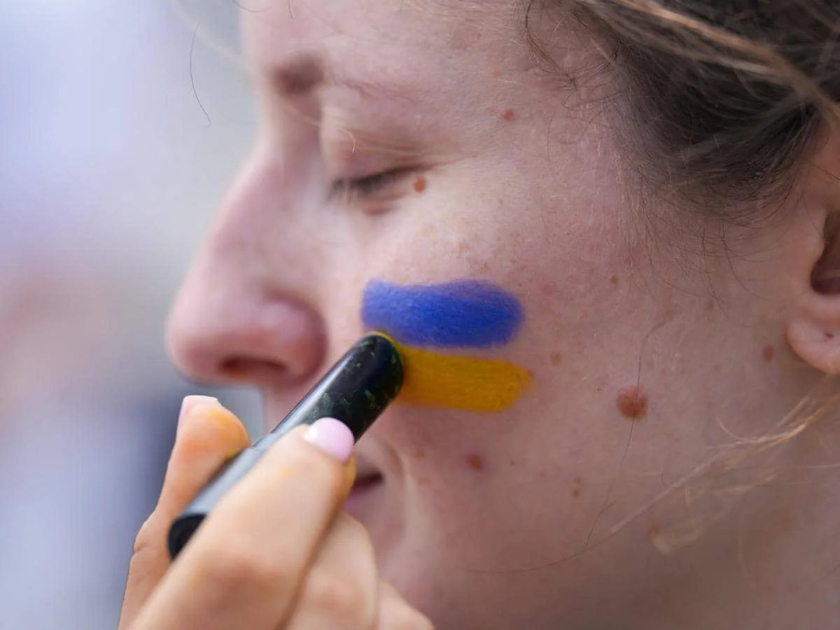 Seorang perempuan menggambar dua garis warna biru dan kuning di pipinya menggunakan facepaint sebagai simbol bendera.
