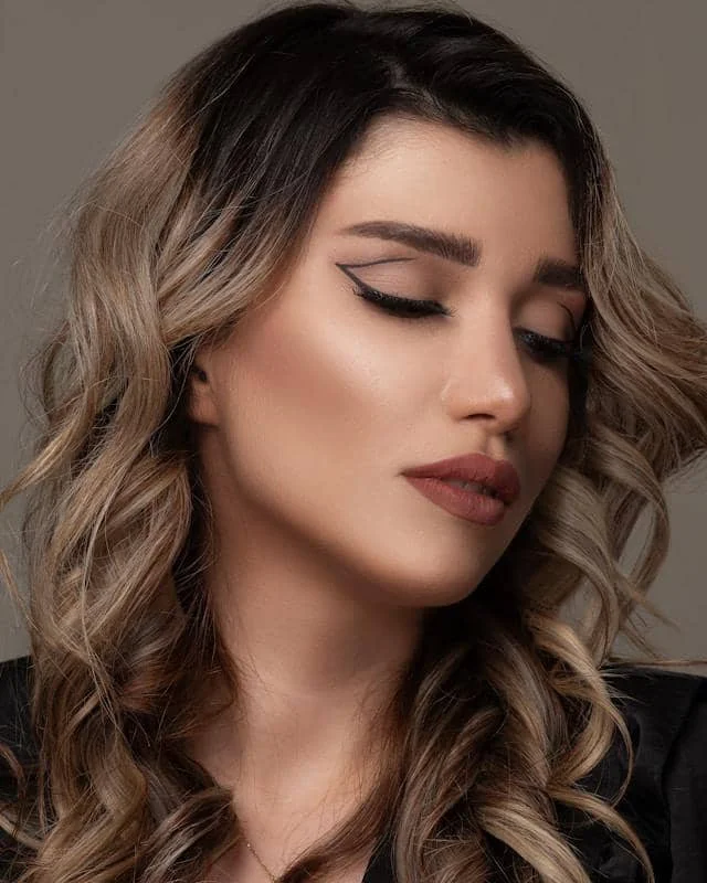 Seorang wanita dengan riasan mata bergaya graphic eyeliner hitam yang tegas, rambut bergelombang pirang, dan bibir berwarna nude matte.