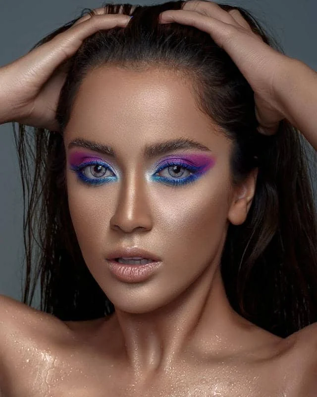 Seorang wanita dengan riasan mata berwarna biru dan ungu mencolok, menatap ke arah kamera dengan tangan di kepala.