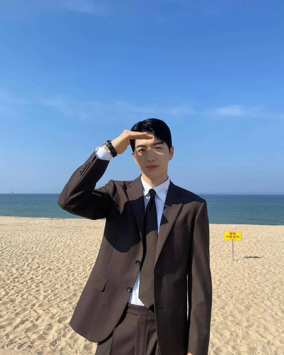 Seorang pria mengenakan setelan jas cokelat dan dasi hitam berdiri di pantai berpasir dengan latar belakang laut biru dan langit cerah.