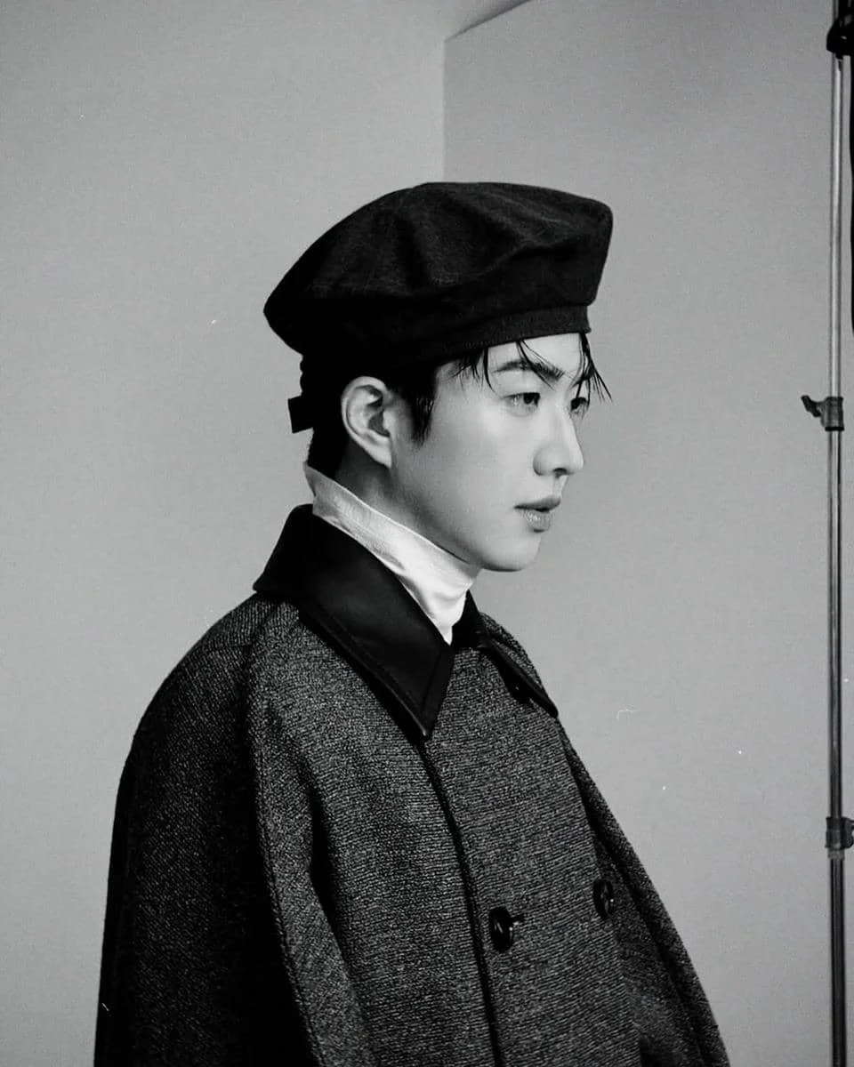 Seorang pria mengenakan mantel tebal dan topi baret hitam berpose dalam foto hitam putih dengan latar sederhana di studio.