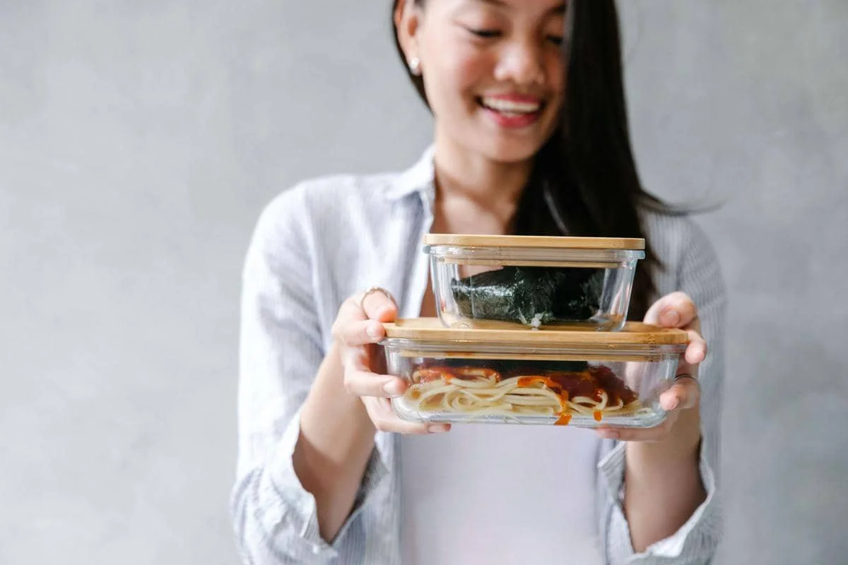 Seorang wanita memegang dua wadah kaca berisi makanan dengan tutup kayu, satu berisi nasi bungkus rumput laut dan satu berisi pasta saus tomat.