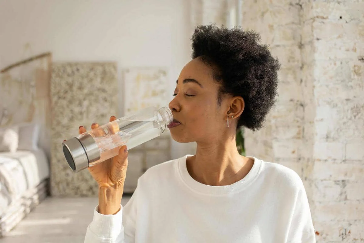 Seorang wanita mengenakan sweter putih sedang minum air dari botol kaca di ruangan terang dengan dinding putih.