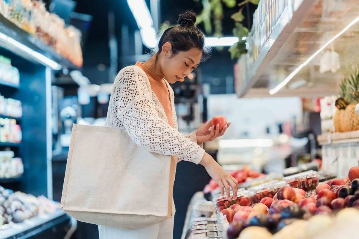 Seorang wanita berbelanja buah segar di supermarket sambil membawa tas belanja kain berwarna krem di bahunya.