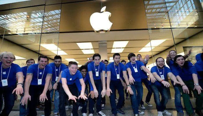 Sekelompok karyawan Apple mengenakan kaus biru berdiri di depan toko Apple dengan logo besar berwarna putih di atas pintu kaca.