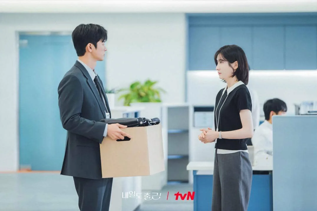 Dua karyawan berdiri saling berhadapan di kantor modern, pria memegang kotak kardus sementara wanita menatapnya serius.