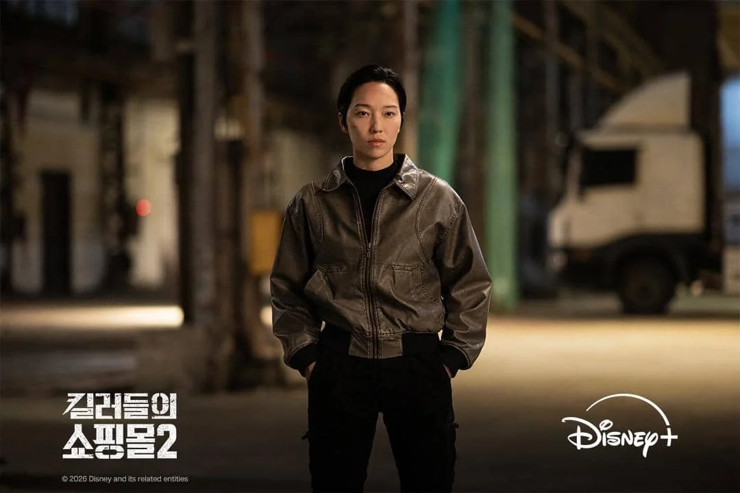 Seorang karakter berdiri di area gudang dengan pencahayaan redup, mengenakan jaket kulit gelap dalam adegan serial A Shop for Killers 2 di Disney+.