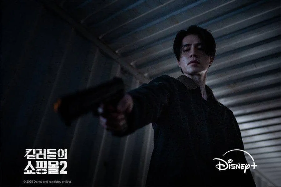 Seorang pria berwajah serius mengarahkan pistol di dalam ruangan logam gelap dalam adegan serial A Shop for Killers 2 di Disney+.