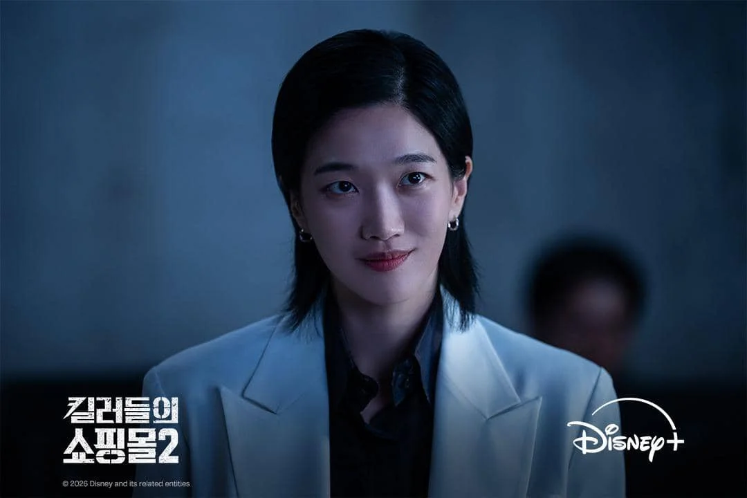 Seorang perempuan berambut pendek mengenakan jas putih dan kemeja hitam tampak tersenyum dalam adegan serial A Shop for Killers 2 di Disney+.