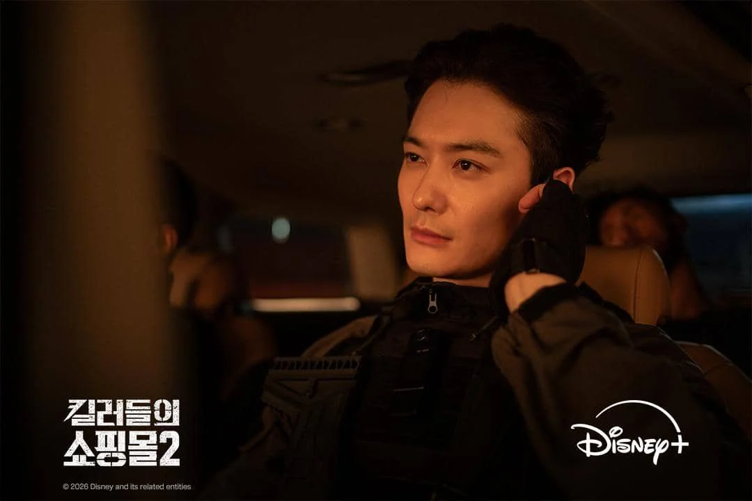 Seorang pria mengenakan pakaian taktis duduk di dalam mobil dengan ekspresi serius dalam adegan serial A Shop for Killers 2 di Disney+.