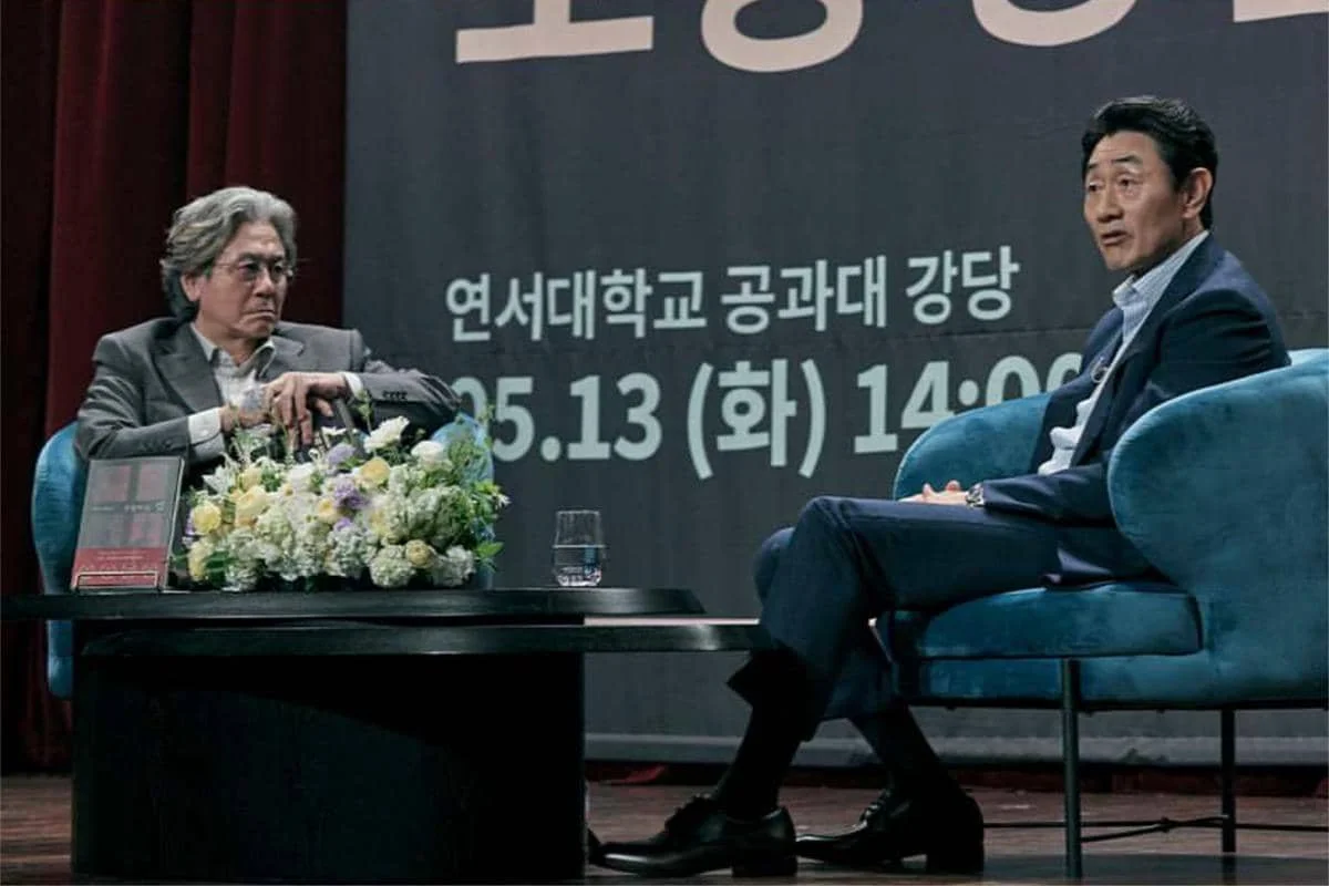 Dua pria duduk di atas panggung saat acara diskusi, dengan meja berisi bunga dan latar bertuliskan teks Korea di belakang mereka.