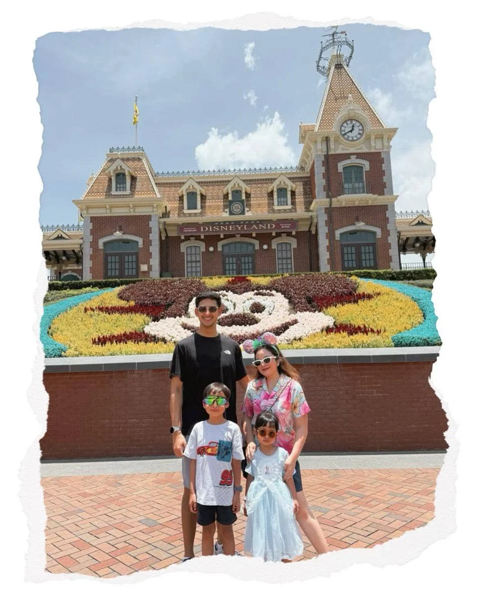 Sebuah keluarga berpose di depan gerbang utama Disneyland Hong Kong dengan latar bunga berbentuk karakter Disney dan bangunan bergaya klasik.