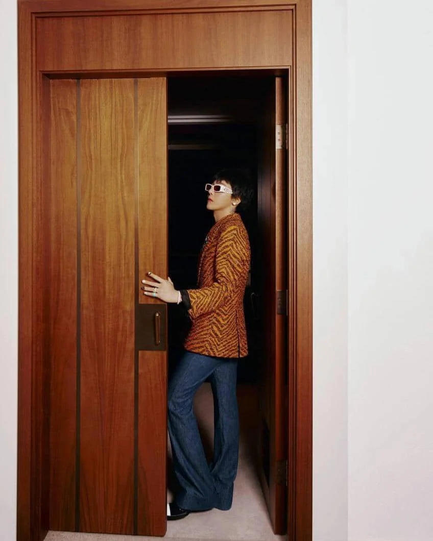 Seorang pria bergaya retro mengenakan jaket bermotif oranye, celana denim lebar, dan kacamata putih berdiri di depan pintu kayu.