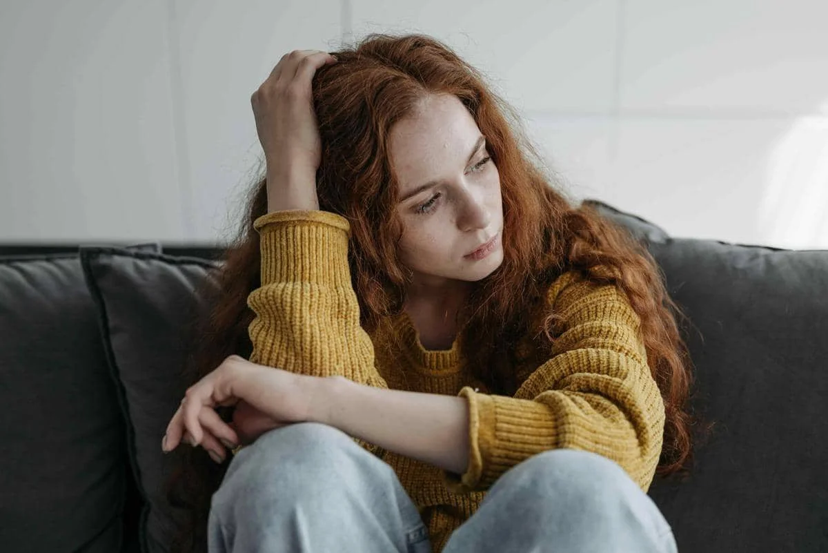 Seorang perempuan berambut panjang duduk di sofa dengan ekspresi murung sambil memegang kepala, mengenakan sweter kuning dan celana jeans.