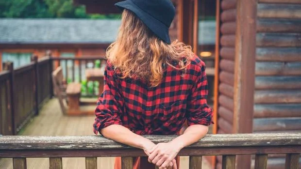 A young woman wearing a hat is relaxing on the porch of a log cabin
