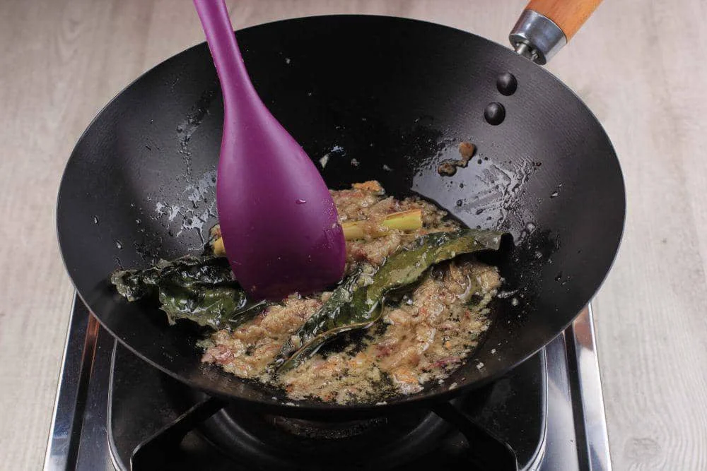 Proses menumis bumbu halus dengan daun jeruk dan serai di wajan hitam menggunakan spatula ungu di atas kompor.