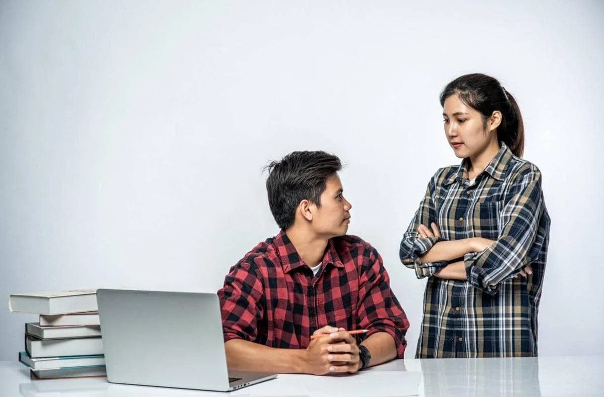 Seorang pria duduk di depan laptop menatap wanita yang berdiri dengan tangan terlipat, menggambarkan situasi tegang atau saling menghakimi.