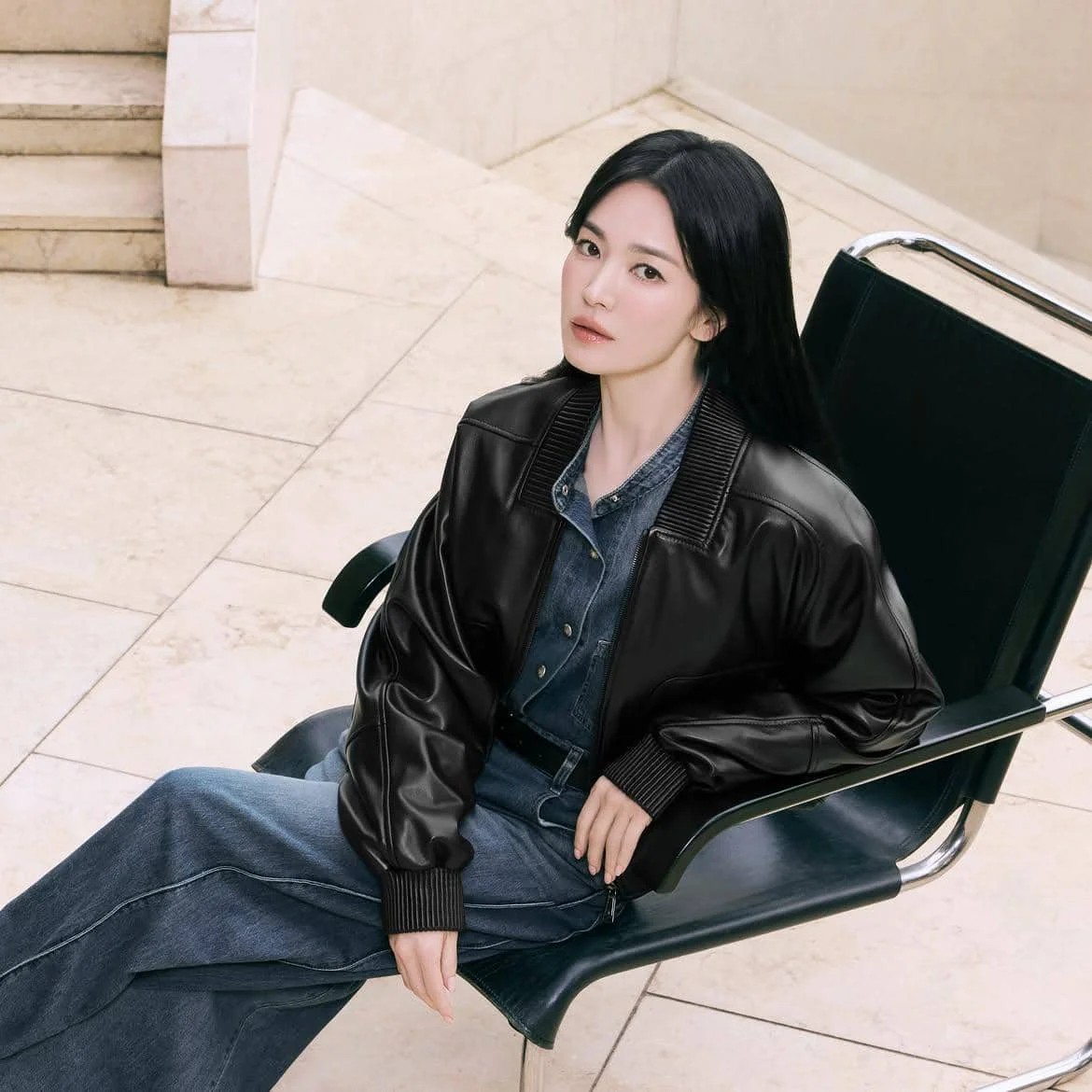 Seorang wanita berambut panjang mengenakan jaket kulit hitam dan pakaian denim duduk di kursi hitam di ruangan bernuansa krem.