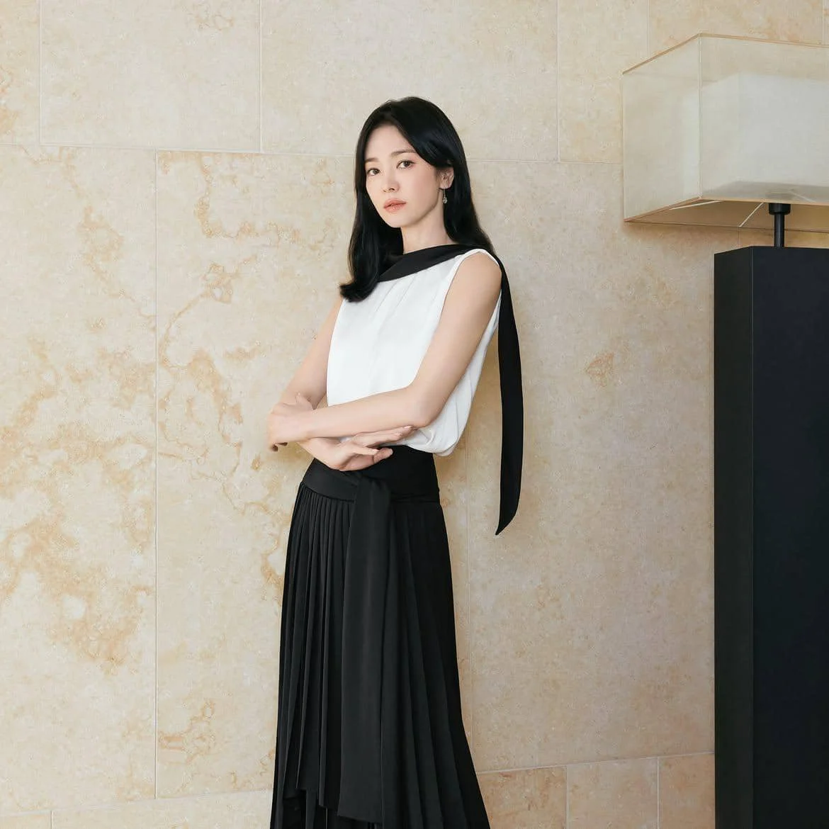 Seorang wanita berambut hitam mengenakan blus tanpa lengan putih dengan detail pita hitam dan rok panjang hitam berdiri di depan dinding krem.