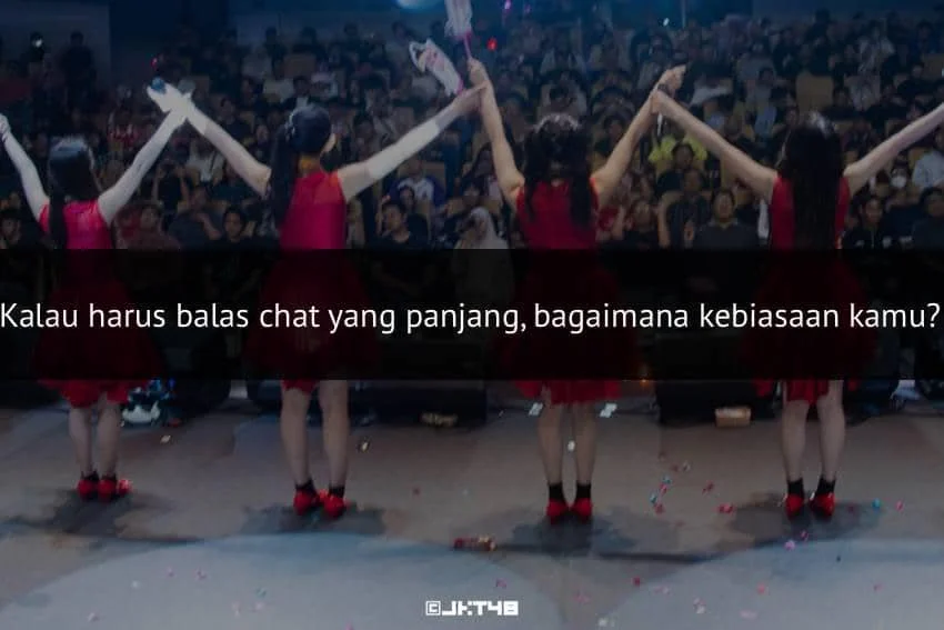 Empat anggota grup idola mengenakan gaun merah berdiri di atas panggung sambil bergandengan tangan menghadap penonton yang memenuhi ruangan.