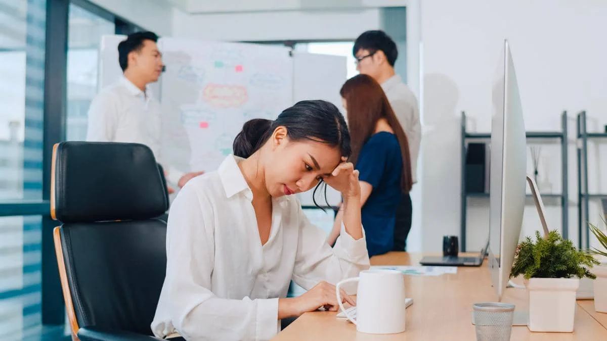 Seorang wanita tampak stres dan berpikir keras di depan komputer di kantor, sementara rekan kerjanya berdiskusi di latar belakang.