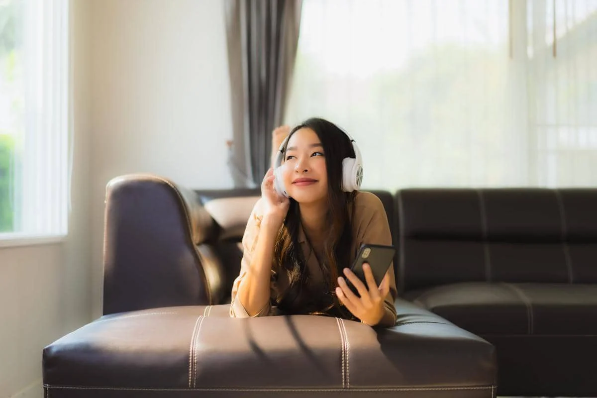 Seorang perempuan berbaring santai di sofa sambil mengenakan headphone putih dan memegang ponsel di ruang tamu yang terang.