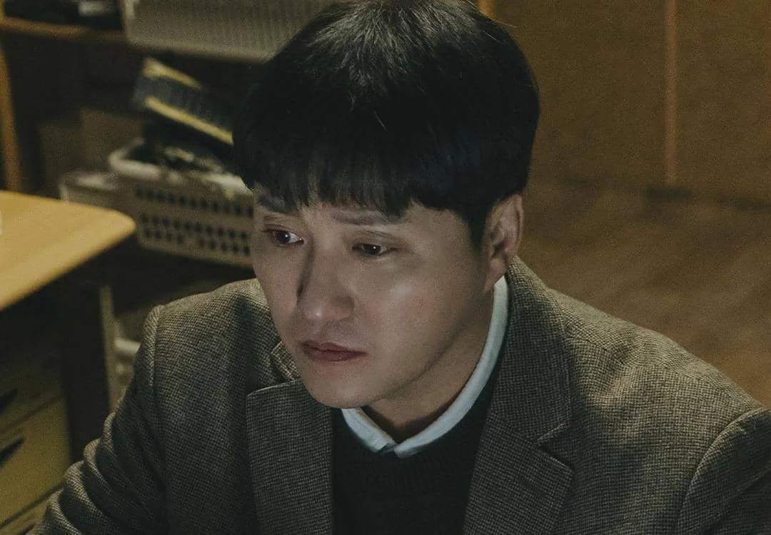 Seorang pria berwajah sedih mengenakan jas cokelat dan kemeja putih duduk di ruangan redup dalam adegan drama Korea The Husband.