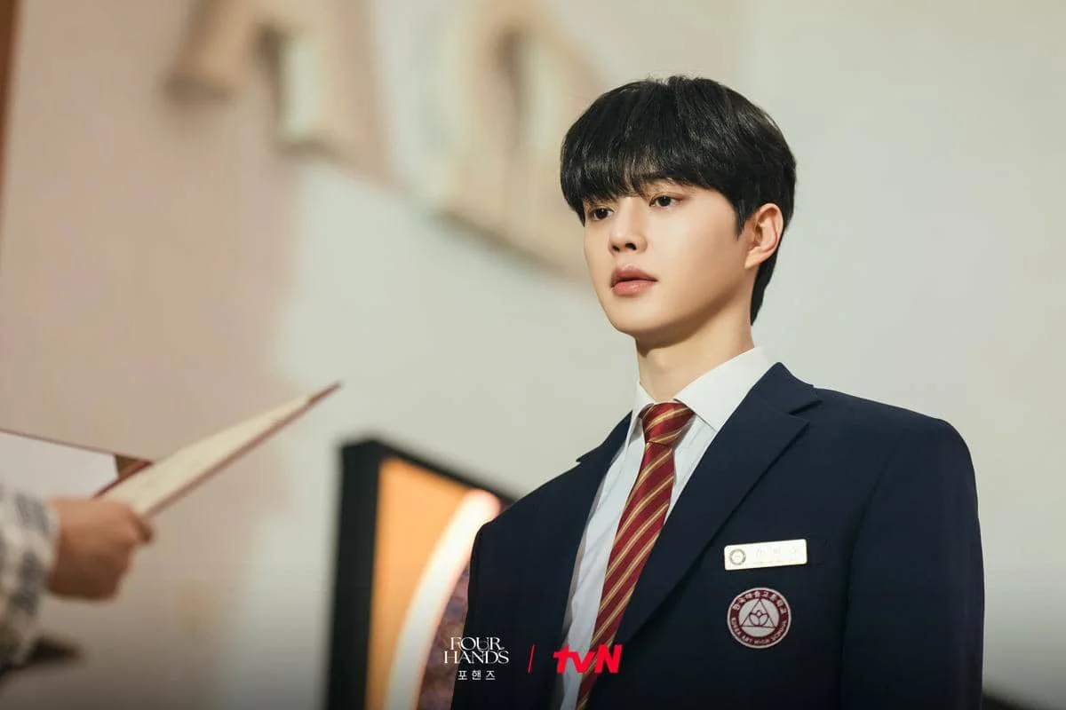 Seorang siswa berseragam sekolah berdasi merah bergaris berdiri serius di dalam ruangan dalam adegan drama Four Hands, Two Sonatas.