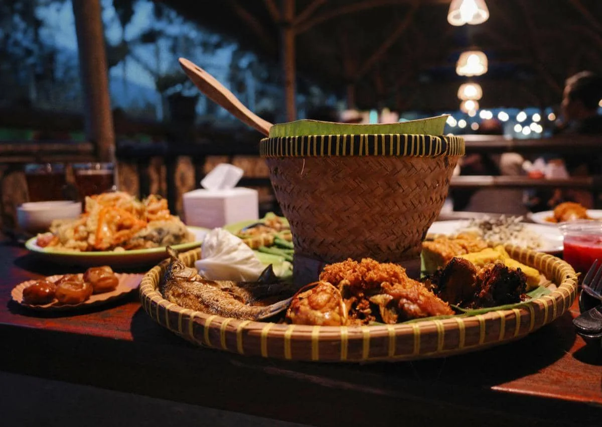 Hidangan tradisional Indonesia tersaji di meja warung makan dengan nasi dalam bakul anyaman dan lauk pauk seperti ayam goreng dan ikan.