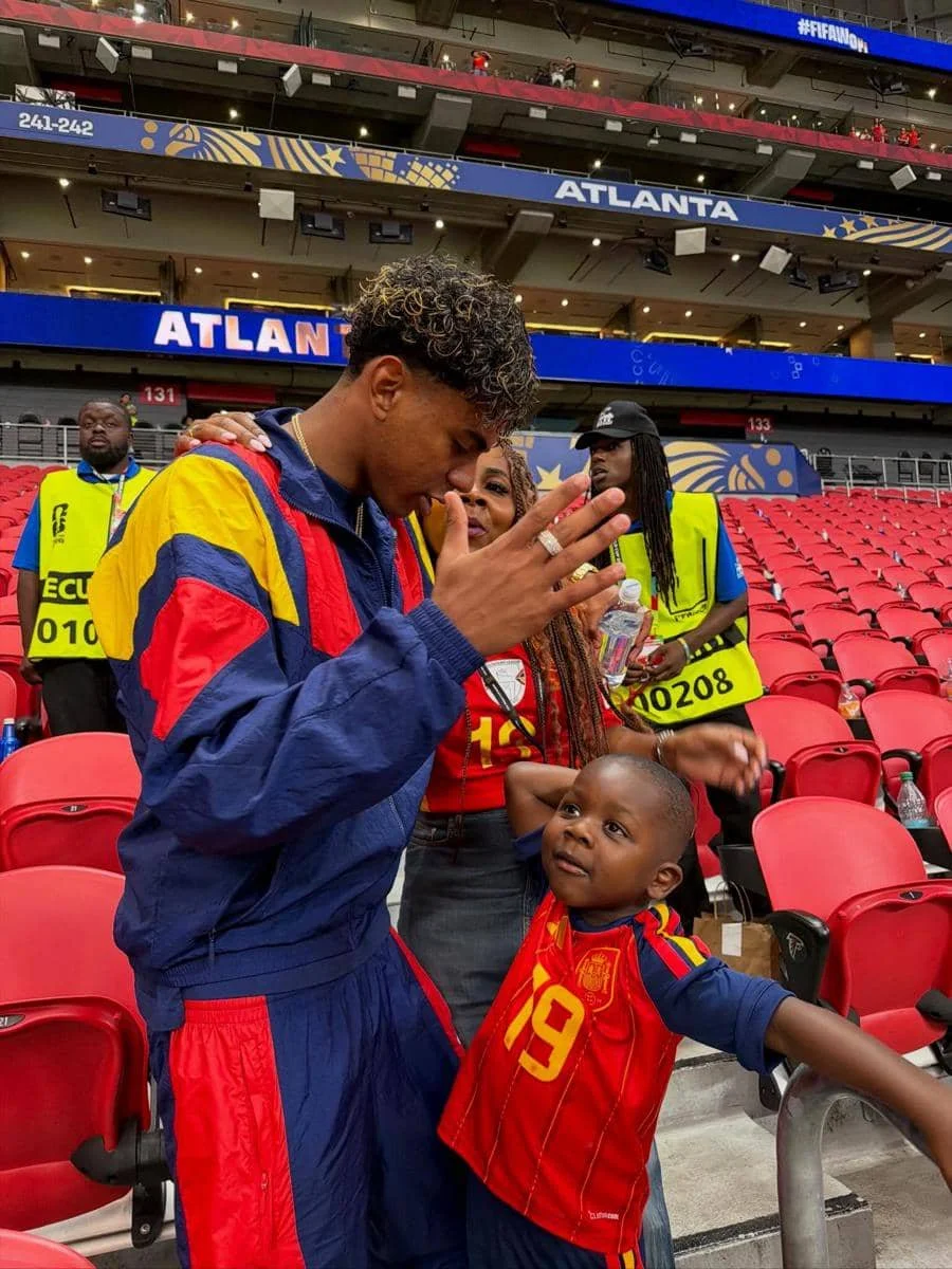Seorang pria mengenakan jaket berwarna merah dan biru berbicara dengan seorang anak kecil yang memakai jersey tim Spanyol di stadion Atlanta.